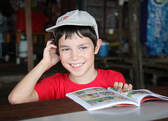 Child in a ball cap smiling at something off-screen. Child appears to be in the middle of reading a graphic novel.