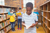 One school-age child is standing among bookshelves, looking at an open book. In the background are three additional children doing the same thing.
