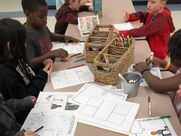 A group of children sit around a long table, coloring. In the middle of the table is a basket of crayons. Worksheets are strewn across the table.