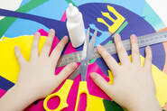 Bird's Eye view of two hands on a child who is out of the frame. The hands lay on scissors, a ruler, and colorful paper. A bottle of glue stands in the middle.