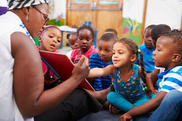 A large group of children gathers around a caregiver who is reading aloud. The caregiver is showing a picture book to the children. One child is pointing at the book.
